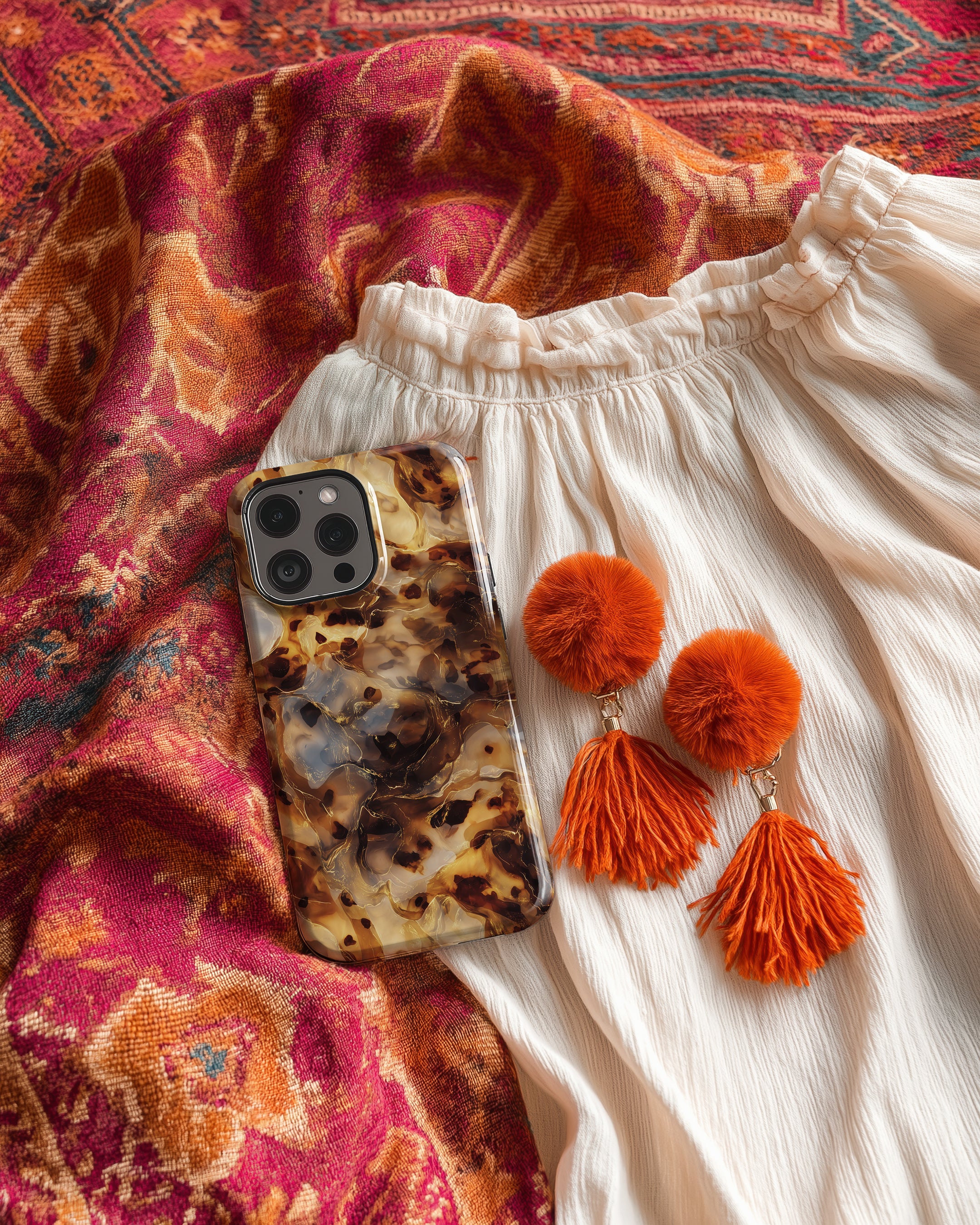 Phone with tortoiseshell case, white pants, and orange pom-pom earrings on a patterned fabric background